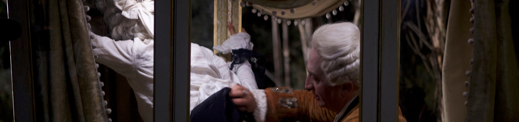 A man in a white wig and 18th century costume lifting up the skirt of a woman who is facing away. She is waring a similar wig. The image is a cropped version of a film still of Théodora Marcadé and Marc Susini in Albert Serra's LIBERTE. Original image Courtesy of Cinema Guild.